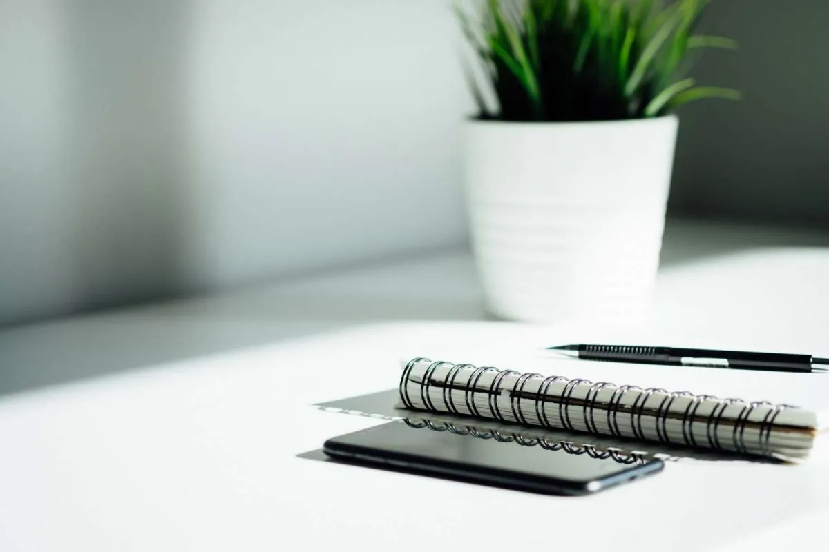 Celular, caderneta, lapiseira e um vaso de plantas de cerâmica branco e pequeno na mesa do escritório de contabilidade