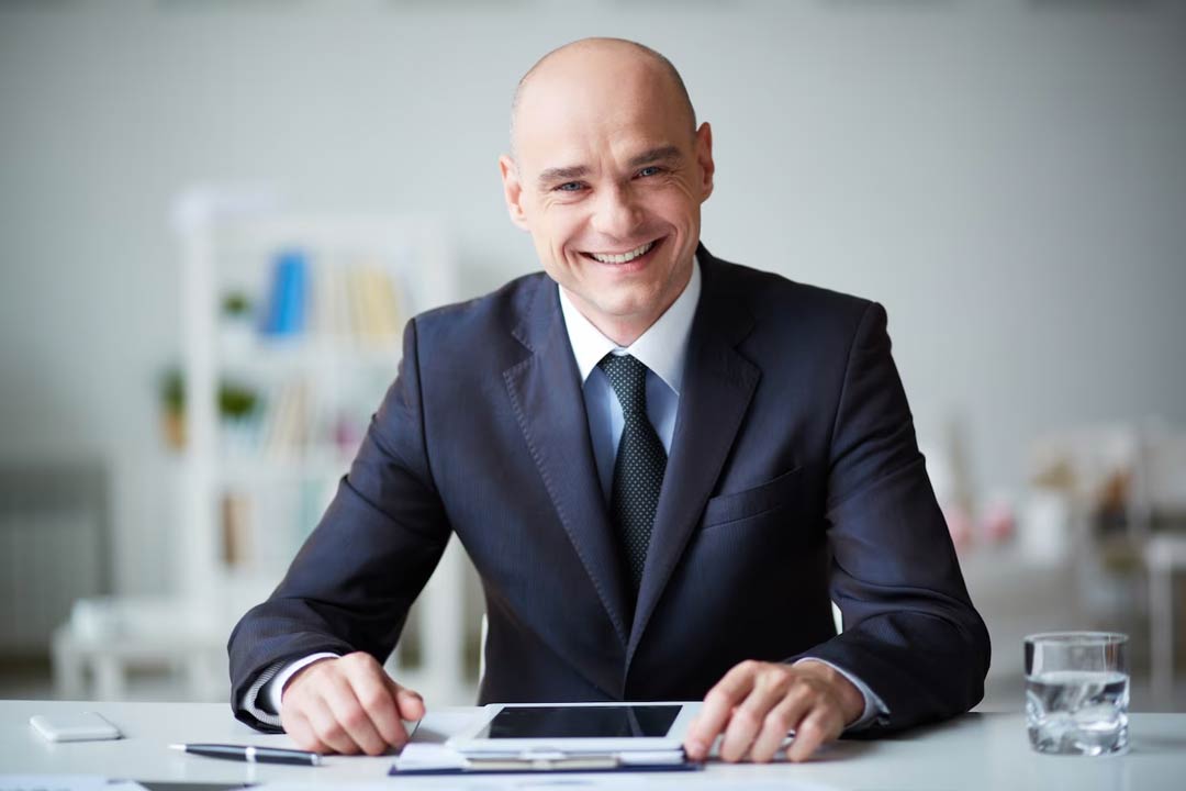 Executivo experiente vestido com terno azul marinho, camisa branca e gravata preta, sentado e sorrindo em mesa de escritório com um copo de água, caneta, celular e tablet