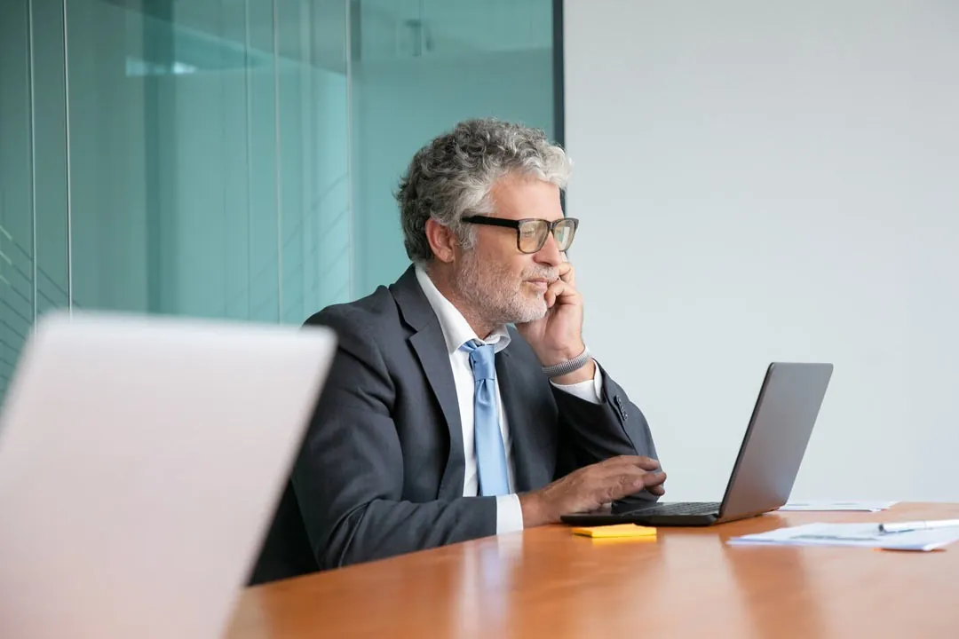 Contador experiente, com os cabelos brancos, óculos de grau, terno e gravata, sentado em mesa de escritório com cara de preocupação analisando dados em um notebook