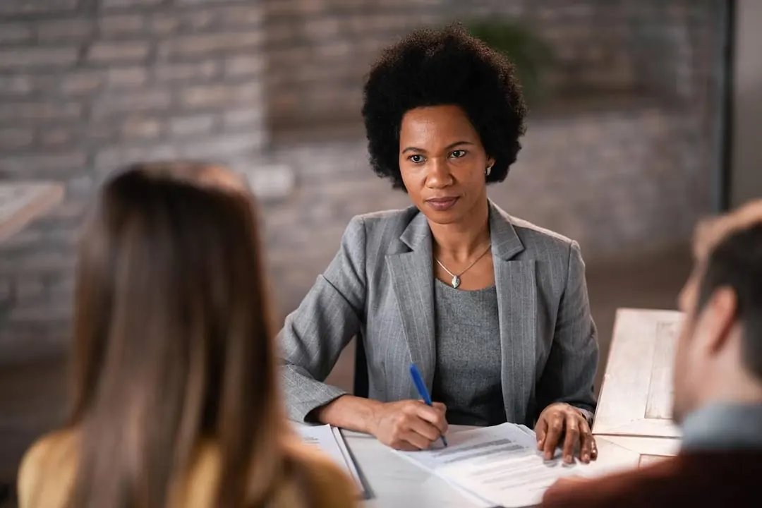 A black executive in a dress suit sits at an office desk interviewing a candidate for a job vacancy