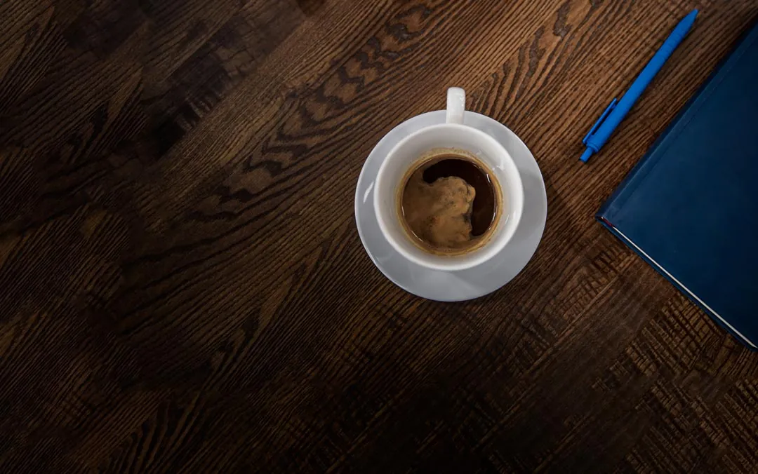 Xícara branca com café, uma caneta azul e uma agenda azul em cima do tampo de madeira de uma mesa de escritório