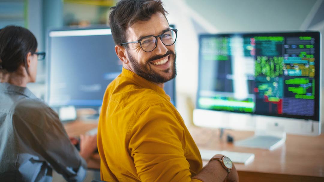 Jovem empresário de óculos, barba e camisa social amarela, sentado em frente ao computador com linguagem de programação, virado para trás, sorrindo