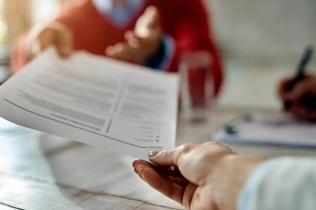 A woman's hand handing a document to a seated person, with a blurred image in the background, at an office desk with another person also with a blurred image making notes on a spreadsheet