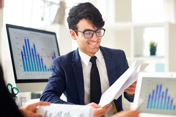 Jovem profissional de contabilidade sorrindo e vestindo roupa social, analisa informações, sobre a empresa, entre as telas de dois computadores com a imagem de gráficos