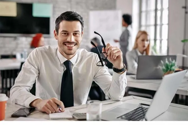 Homem, profissional de contabilidade, sentado em mesa de escritório segurando óculos de grau com armação preta, sorrindo, com colegas de trabalho ao fundo com a imagem desfocada