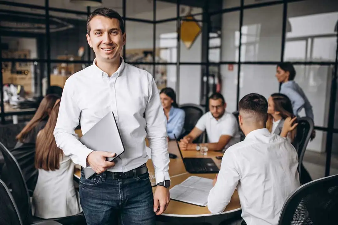Young businessman smiling, holding a notebook in front of a table with a group of businessmen sitting with notebooks and notebooks, in a meeting