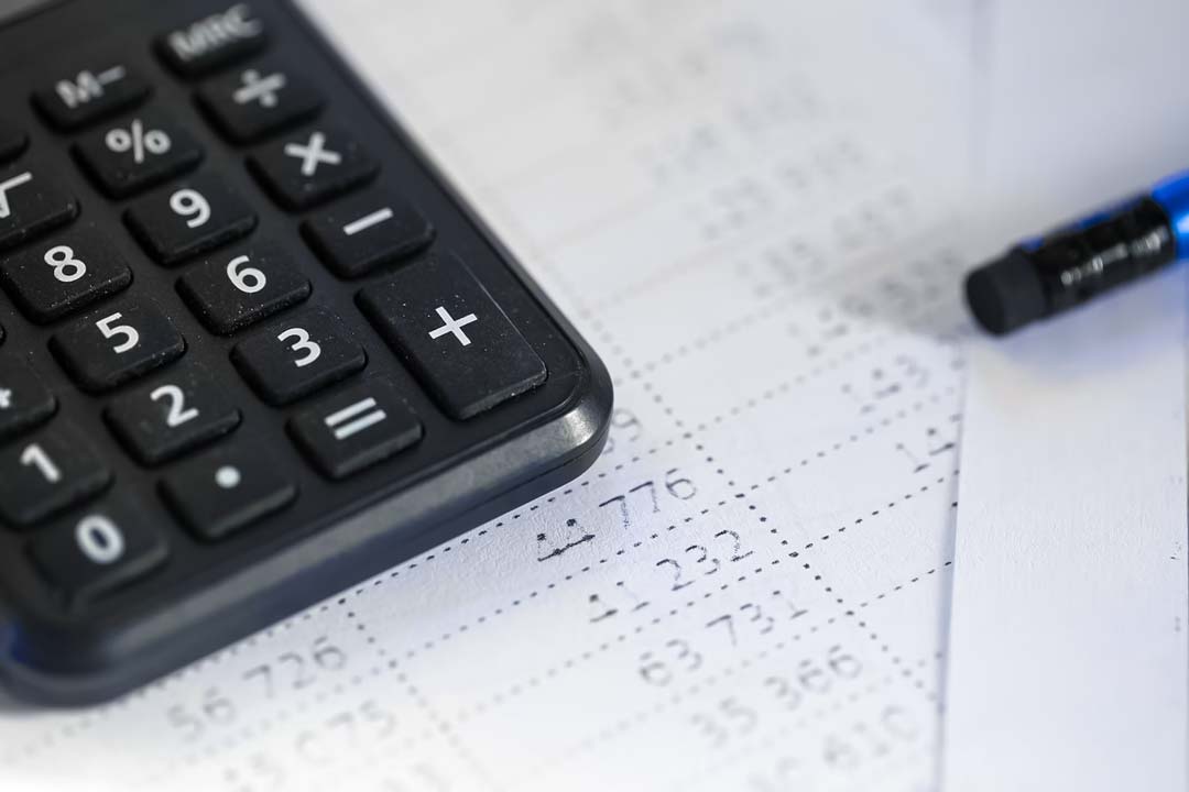 Parte de uma calculadora na cor preta e parte de trás de um lápis azul com ponta de borracha em cima de uma folha de sulfite com informações de uma planilha com números