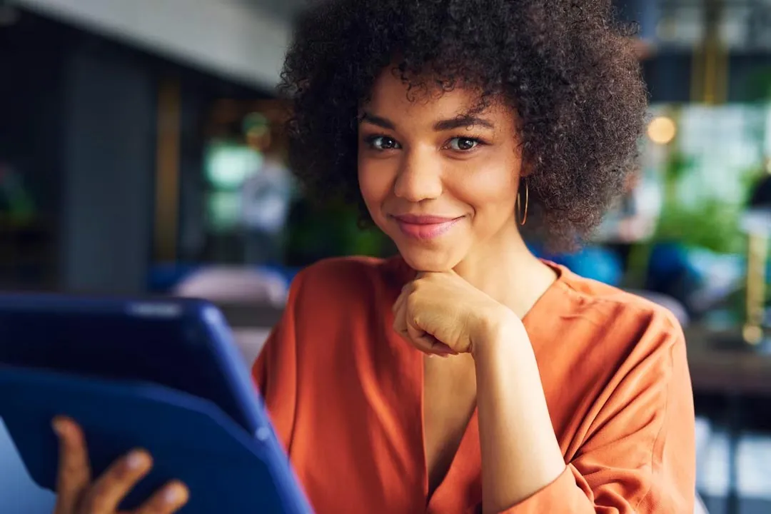 Jovem mulher negra, com blusa cor de laranja, segurando um tablet e sorrindo com a mão no queixo dentro do escritório