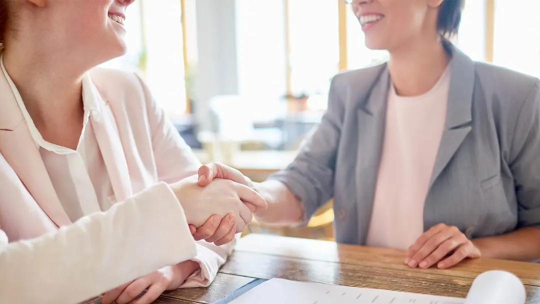 A businesswoman in pink business attire shakes hands with an executive in gray business attire, the two are sitting at an office table, smiling