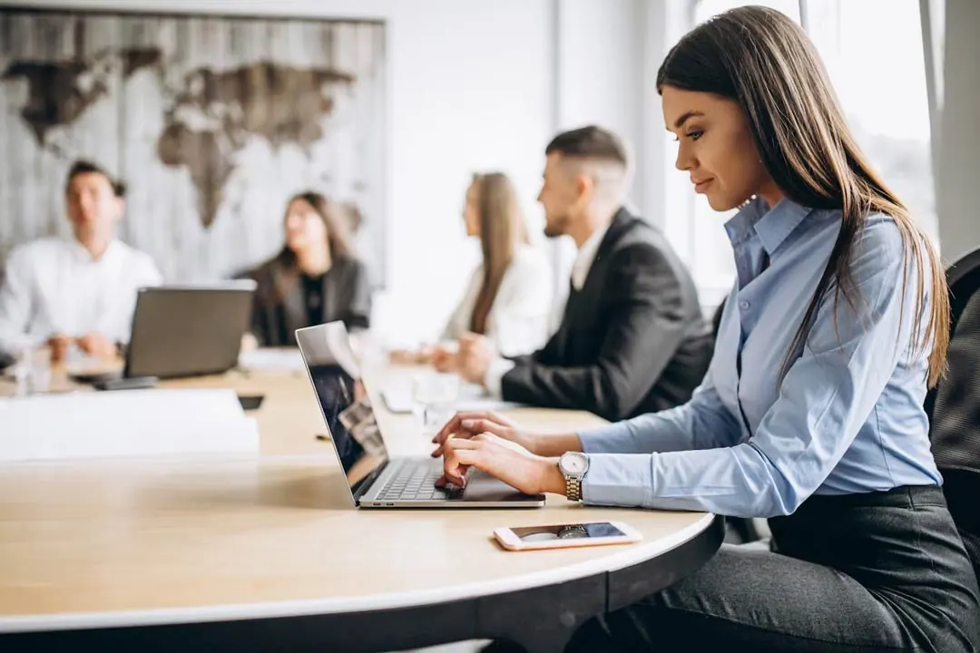 Executiva morena com cabelos lisos compridos, vestida com roupa social feminina, sentada em mesa compartilhada com outras pessoas, dentro de escritório, utilizando um notebook