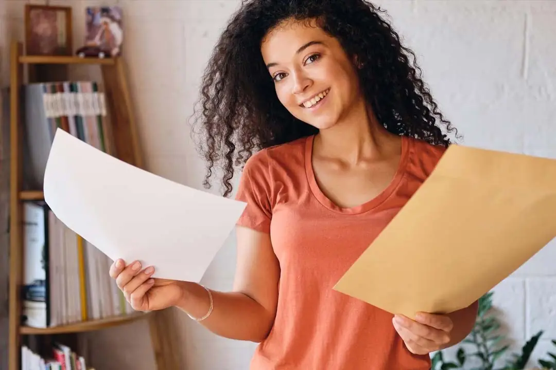 Una joven negra, sonriente, sostiene un sobre naranja en una mano y un documento impreso en papel blanco en la otra