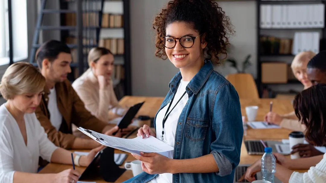 Jovem mulher negra com cabelos presos usando óculos, vestindo uma camiseta branca com uma camisa jeans aberta por cima, sorrindo, segurando uma folha com informações impressas em um ambiente de escritório com outras pessoas desfocadas ao fundo
